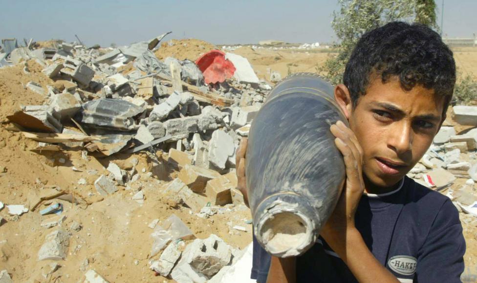 Một cậu bé người Palestine ôm mảnh pháo đứng trước ngôi nhà bị phá hủy bởi cuộc tấn công của lực lượng Irael.