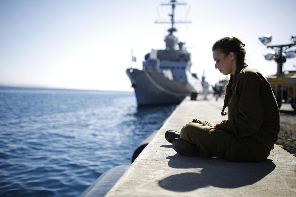 Một nữ binh sĩ Israel ngồi lặng trước biển, gần đó là tàu hải quân đang đậu trên cảng quân sự tại Eilat, Israel. Nguồn ảnh: REUTERS/ Amir Cohen.