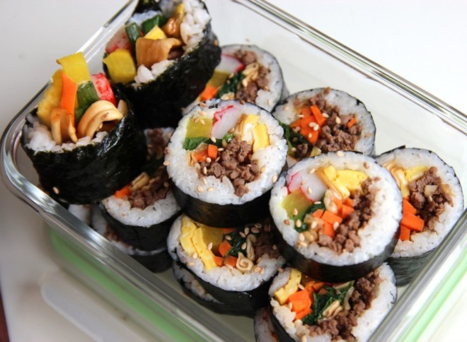 Xếp thứ hai trong danh sách những món ăn nổi tiếng của Hàn Quốc, chắc chắn không còn cái tên nào khác ngoài Kimbap. Kimbap – “kim” là tên ghép của  lá rong biển khô; “bap”  là “cơm”. Tên gọi của món ăn rất đơn giản, cơm gói trong lá rong biển.  Kimbap “có vẻ” giống với Maki – cũng là món cơm cuốn trong lá rong biển của Nhật. Nhưng kimbap thường to hơn (béo hơn) vì bên trong, “nhân” gồm nhiều loại thực phẩm đang dạng hơn gồm cơm trắng, cà rốt, trứng, dưa chuột…, tùy sở thích từng người.