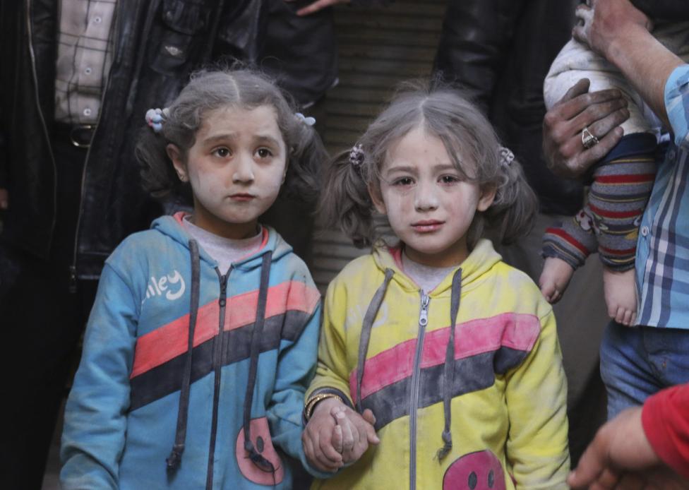 Hai bé gái xinh xắn bị bụi đất phủ đầy người, nắm chặt tay nhau không rời. Hai em đã may mắn sống sót sau một vụ tấn công tên lửa đất đối đất của lực lượng Tổng thống Syria Bashar al-Assad tại huyện Bab al-Hadeed, Aleppo vào tháng 7/2015